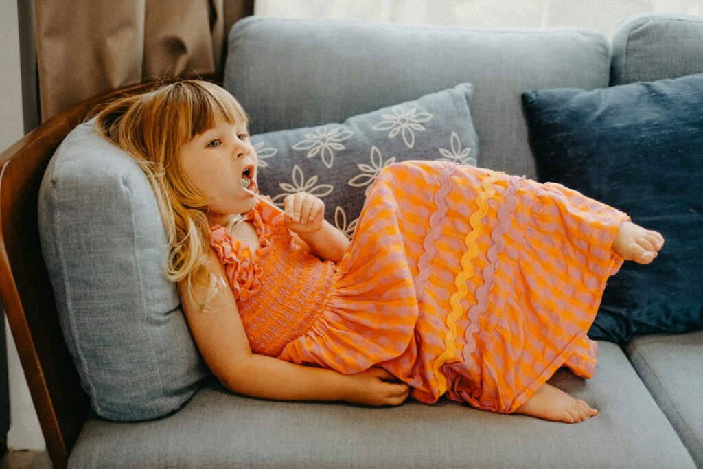 young girl in orange dress sitting on a couch eating lollipop