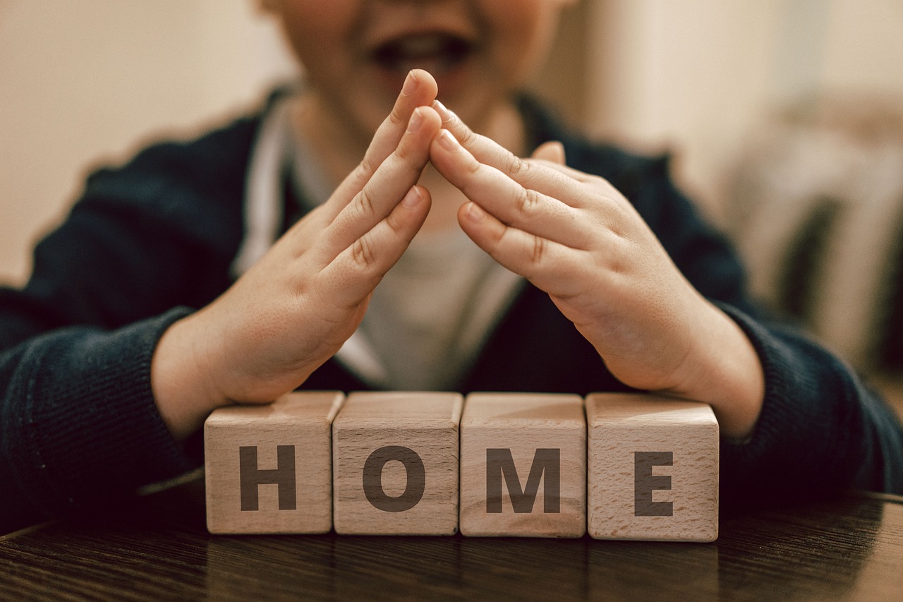 A child with hands gesturing a roof above letter bricks spelled home.