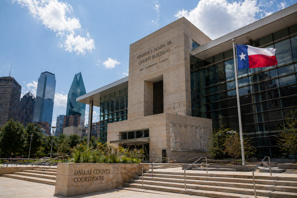The image depicts the exterior of a modern courthouse in Dallas, Texas, showcasing sleek architecture and prominently featuring the Texas flag. This courthouse is a central location for family law matters, including child custody and support cases, as well as divorce proceedings under Texas law.