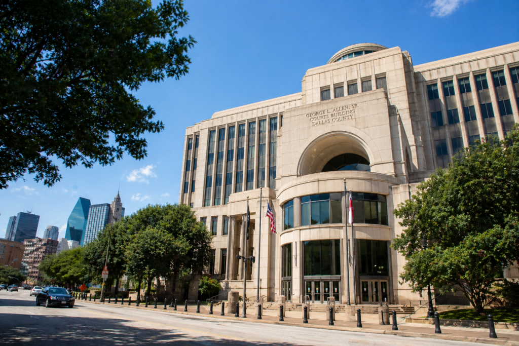 The image shows the exterior of a courthouse in an urban Texas setting, with a clear blue sky and sunlight illuminating the building. This venue is often involved in family law matters, including divorce proceedings and child custody arrangements in Dallas County.
