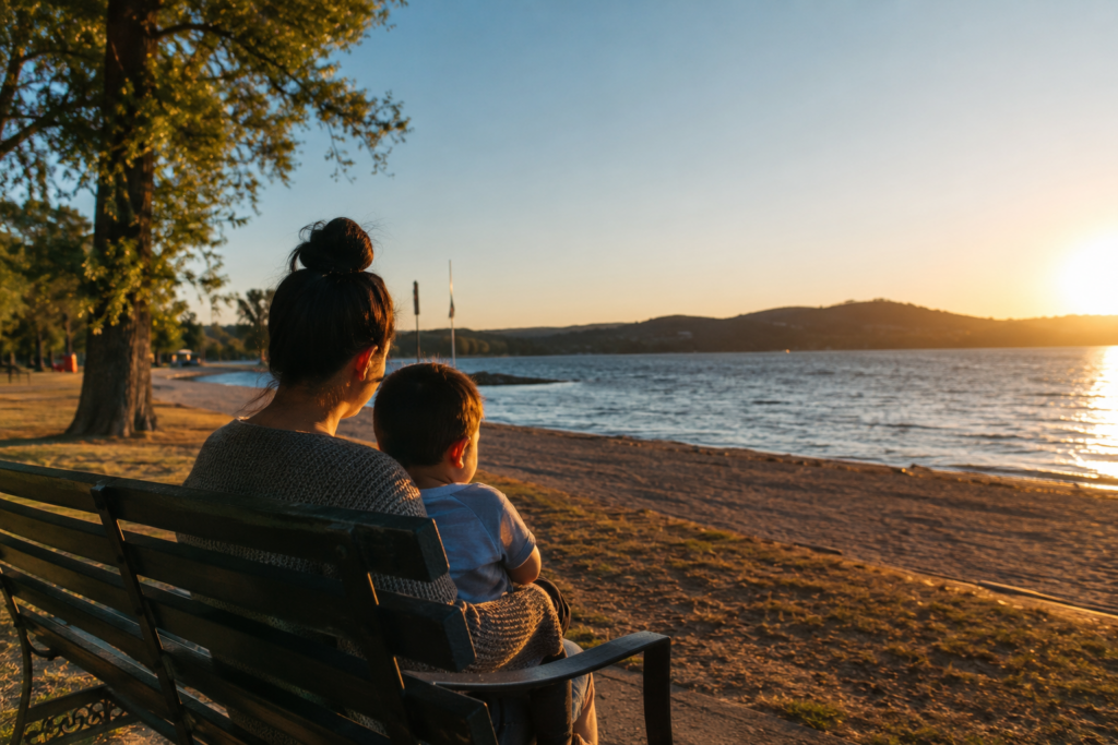 Wide-angle, cinematic summer scene of a parent and child sitting on a bench by a sunlit lake at sunset, evoking reflection, urgency, and the limited time families face in CPS cases.