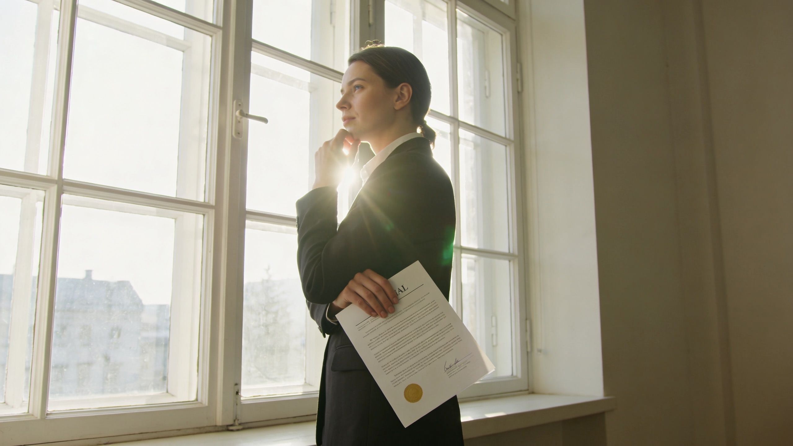 A professional woman in a suit holding a legal document while standing thoughtfully before a bright window.